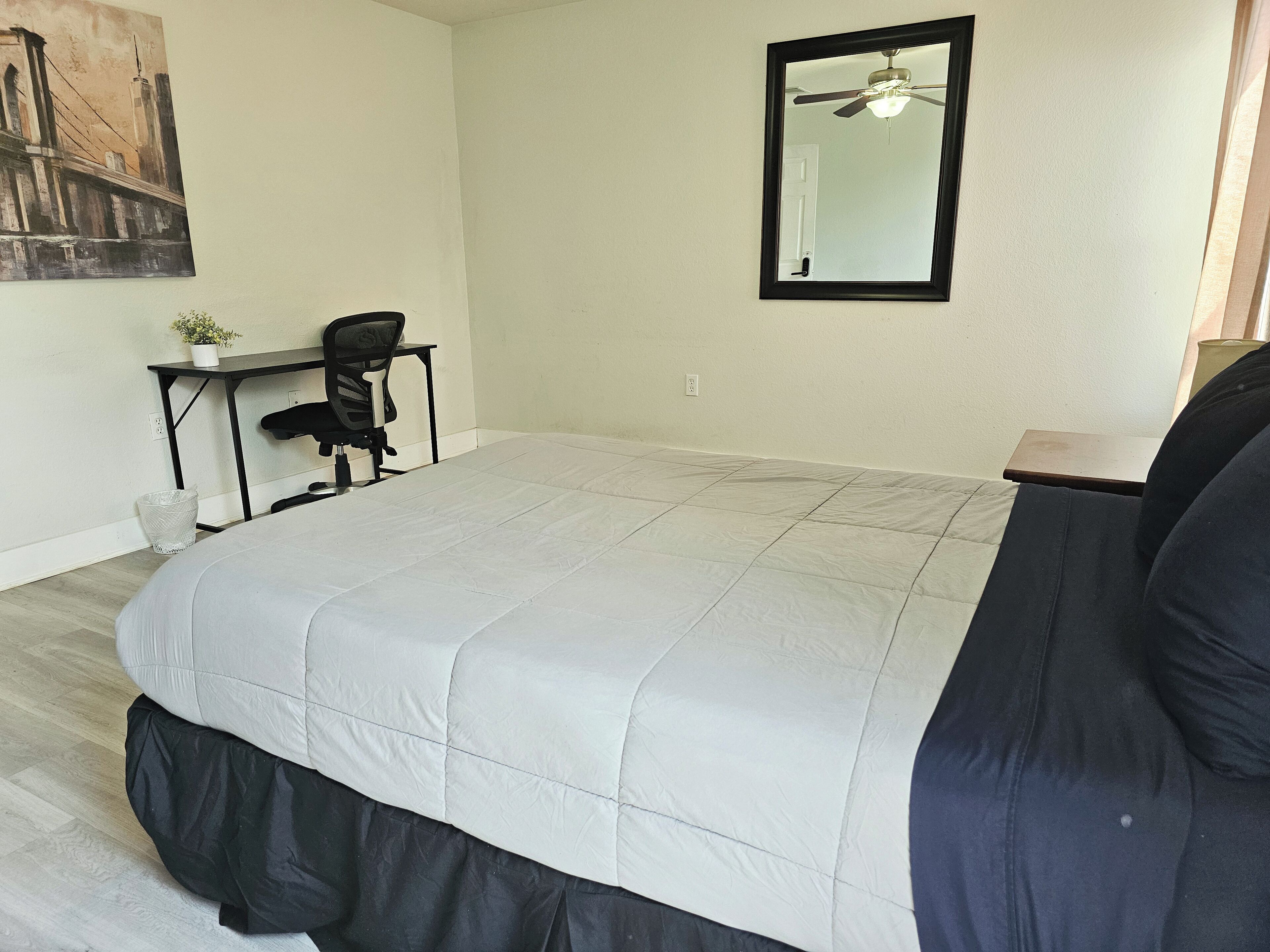 A bedroom with a large bed, a desk with a chair, a ceiling fan, and a painting on the wall.