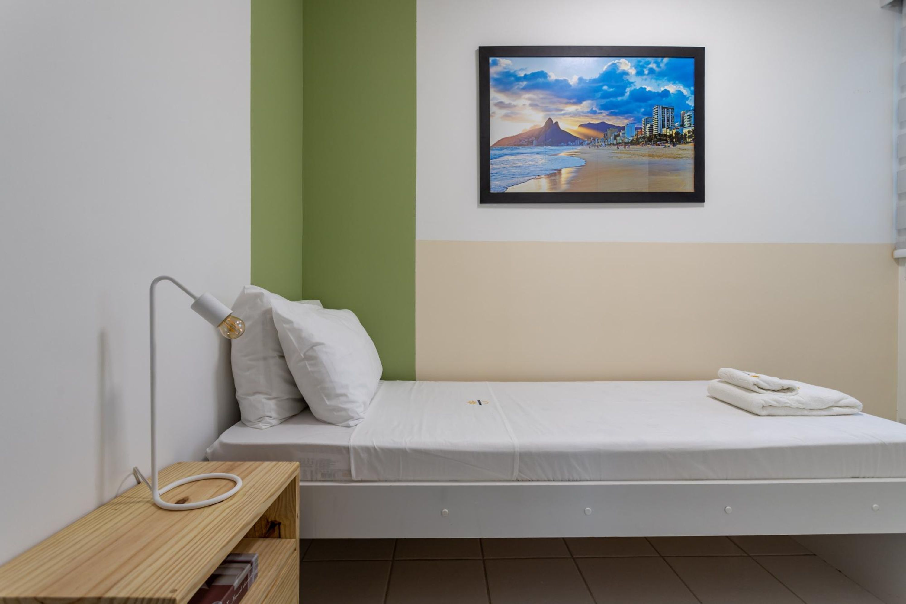 Una cama individual con colchón y almohadas blancas, una mesita de noche con lámpara, y un cuadro con una imagen de una playa en la pared.