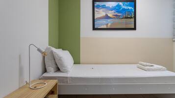 Una cama individual con colchón y almohadas blancas, una mesita de noche con lámpara, y un cuadro con una imagen de una playa en la pared.