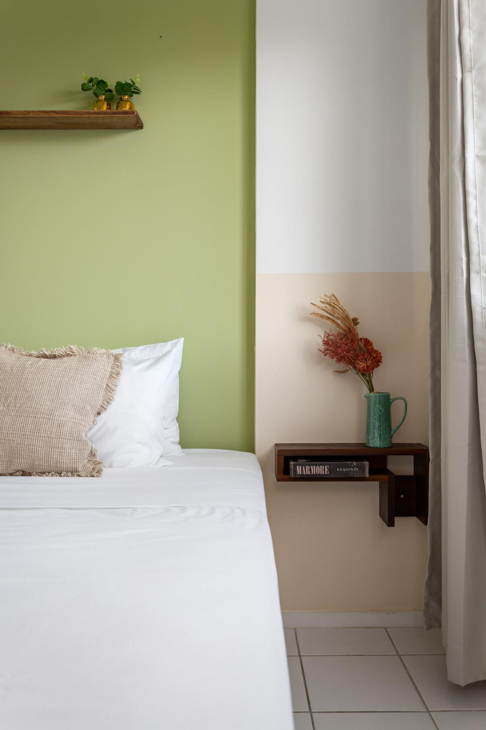 A neatly made bed with white bedding and a beige throw pillow. A small wooden shelf with a vase of dried flowers and a book titled "Marmore" is mounted on the wall.