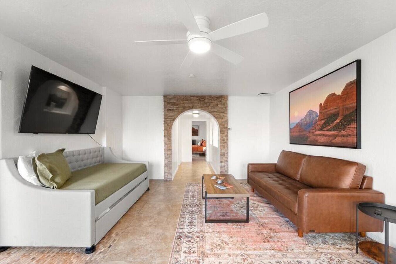 A modern living room with a white sofa, a brown leather couch, a coffee table, and a TV mounted on the wall. There is a brick wall and a painting of a landscape.