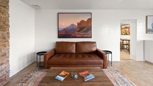 Een woonkamer met een bruine leren bank, een bijzettafel met boeken, een schilderij van een woestijnlandschap en een accentmuur van steen.