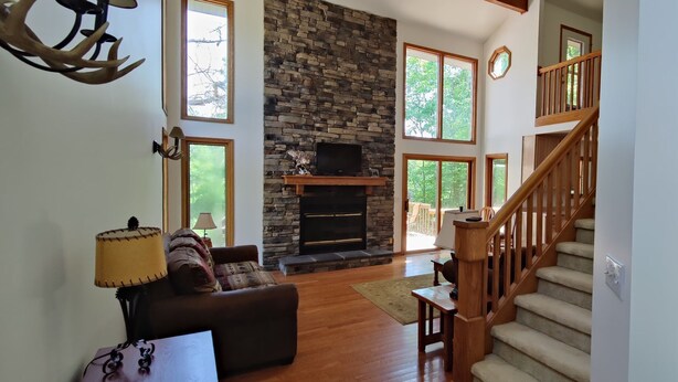 A spacious living room with a stone fireplace, a wooden staircase, and large windows offering a view of the outdoors. - Riverside/Fishing/Arcade/Pool Table/Disk Golf/Fire Pit/Basketball/Pets/WiFi/UTV (Marion)