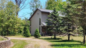 A two-story wooden cabin with a sloped roof, surrounded by a gravel path and lush greenery. - Riverside/Fishing/Arcade/Pool Table/Disk Golf/Fire Pit/Basketball/Pets/WiFi/UTV (Marion)