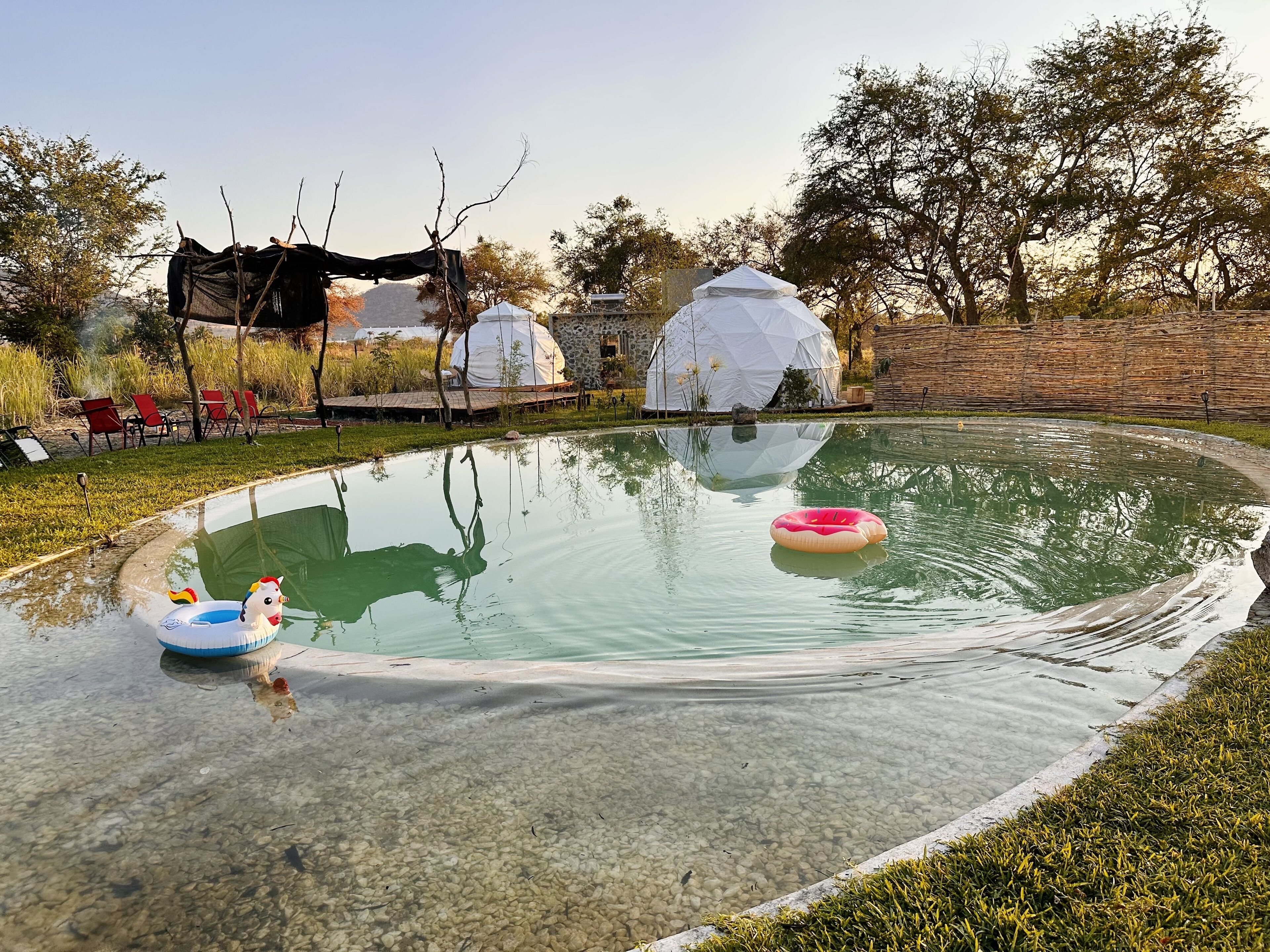 Una piscina con acqua cristallina, circondata da un'area verdeggiante e alberi. Sullo sfondo, si notano due strutture bianche a forma di cupola.