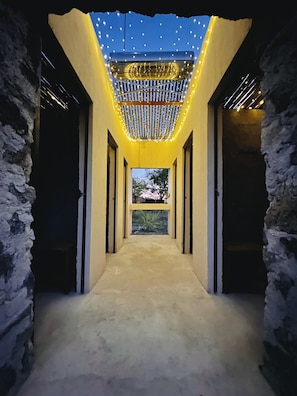 A corridor with a ceiling adorned with string lights, flanked by stone walls and leading to a window with a view of trees.