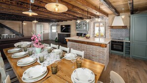 Un comedor con una mesa de madera dispuesta para seis comensales, una cocina con salpicadero de piedra y vigas de madera a la vista en el techo.