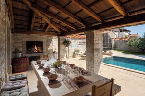 A stone-walled dining area with a long table set for a meal, a fireplace, and a poolside view.