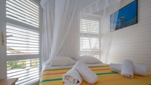 Een hotelkamer met een bed, witte gordijnen, een schilderij en uitzicht op groen.