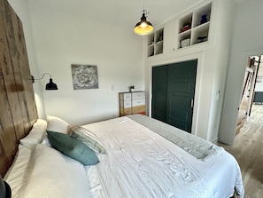 A bedroom with a bed, pillows, a dresser, a door, and a wall-mounted lamp.