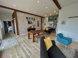 A modern living room with a wooden dining table, a sofa, and a blue armchair. There is a kitchen in the background with a green backsplash.