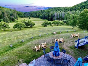 A patio area with outdoor furniture and potted plants, surrounded by a grassy lawn and trees, with a view of a forested hill in the background. - Auberge Val Carroll (Harrington)
