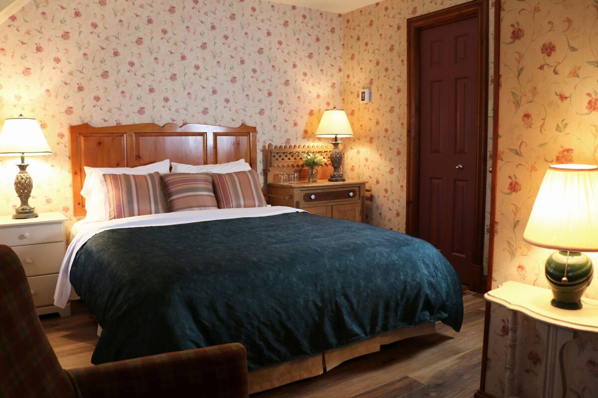 A bedroom with a bed, two bedside tables, a nightstand, a door, and floral wallpaper.