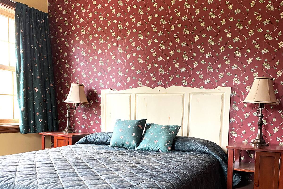A bedroom with a large bed, floral wallpaper, two bedside tables with lamps, and a window with curtains.