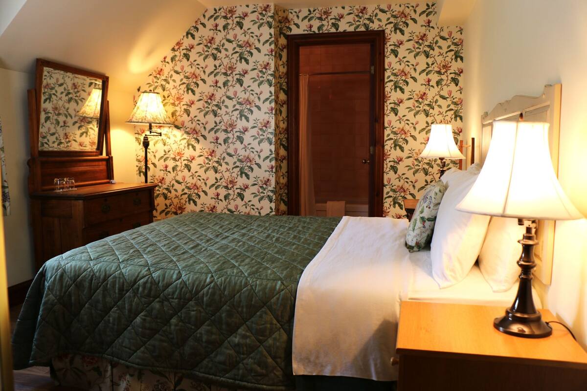 A bedroom with a green quilted bedspread, two bedside lamps, a wooden nightstand, a mirror, and floral wallpaper.
