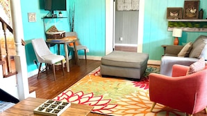 Uma sala de estar com piso de madeira, ventilador de teto, um sofá, uma poltrona, uma mesinha e um tapete colorido.