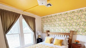 Quarto com cama de madeira, colcha xadrez amarela e branca, papel de parede estampado com flores e janela com cortinas.