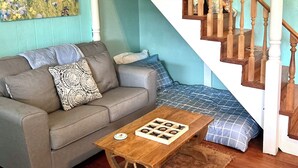 Uma sala de estar com um sofá cinzento, uma mesa de centro de madeira, uma cama com padrão azul e branco, e uma escadaria de madeira.