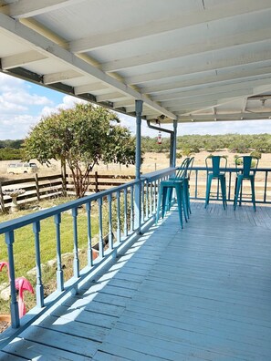 A covered outdoor seating area with blue chairs and a wooden deck, overlooking a fenced field with trees and a distant vehicle. - Escape the city Life,  sit on the Big Porch at Farmhouse with open pasture views (Spring Branch)