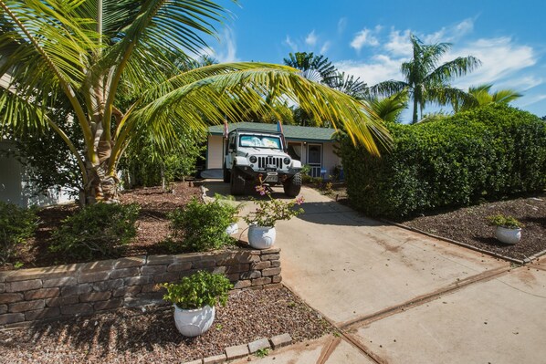 A white off-road vehicle parked in front of a house with a paved driveway, surrounded by palm trees and shrubs.