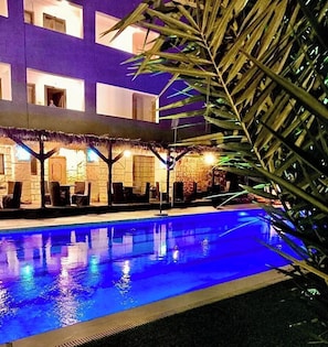 A pool area with a thatched roof seating area, a pool with blue lighting, and a building with multiple windows.