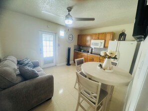 A living room with a sofa, a dining table with chairs, a ceiling fan, and a kitchen with wooden cabinets and a microwave.