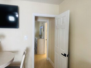 A hallway with a white door, a flat-screen TV mounted on the wall, and a bathroom with a toilet and sink.