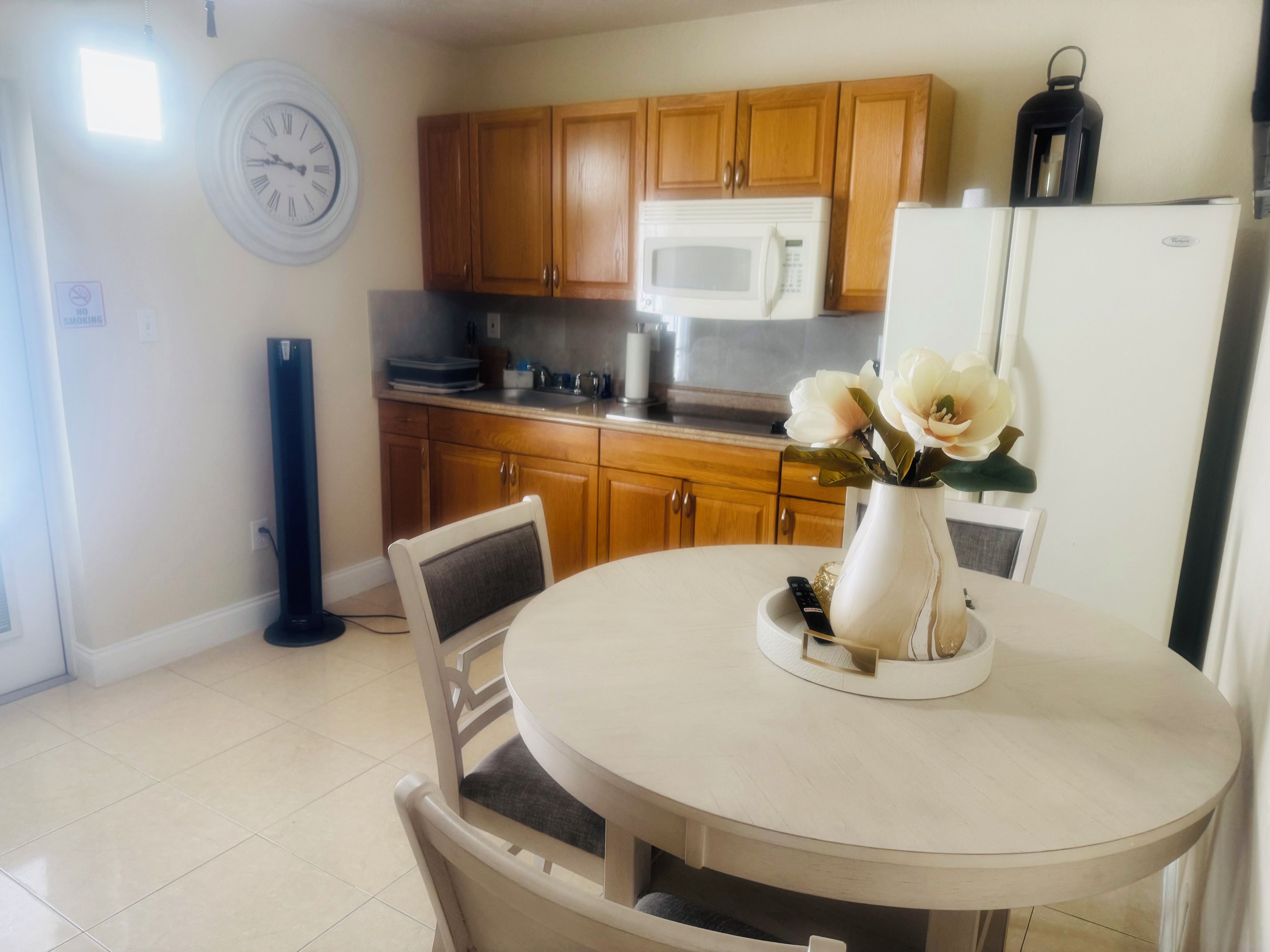 Una cocina con armarios de madera, microondas, refrigerador, mesa de comedor con sillas y un florero con flores.