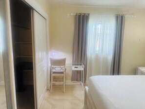 Quarto com cama, cadeira, mesinha, guarda-roupa e janela com cortinas.