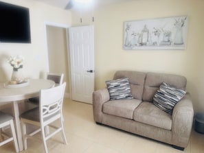 Un salon avec un canapé beige, deux coussins à motifs, une table à manger blanche avec des chaises et un téléviseur à écran plat fixé au mur.