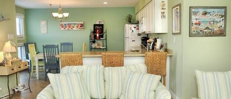 A cozy living room with a striped sofa, a dining area with chairs and a table, and a kitchen with white cabinets and a refrigerator.