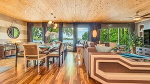 Ein geräumiger Wohnbereich mit Holzboden, ein Essbereich mit Stühlen und einem Tisch sowie ein großes Fenster mit Blick ins Freie.
