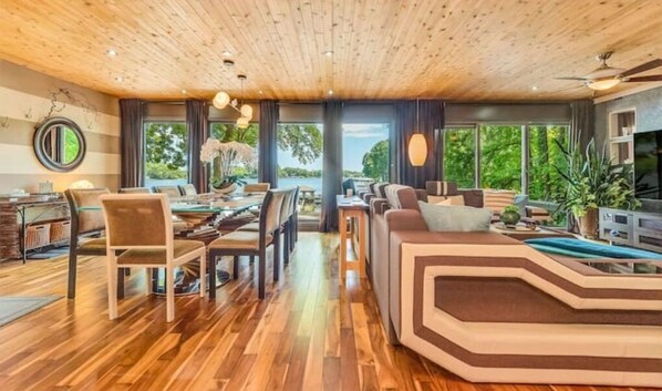 A spacious living area with a wooden ceiling, a dining area with chairs and a table, and a large window offering a view of the outdoors.