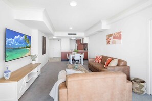 A modern living room with a flat-screen TV, a sofa, a coffee table, and a dining area in the background. - Chevron Renaissance River View (Surfers Paradise)