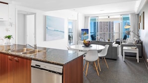 A modern kitchen with a granite countertop, stainless steel sink, and a dishwasher. There is a dining area with a round table and chairs, and a sofa. The room has large windows with a view of buildings. - Chevron Renaissance Ocean View (Surfers Paradise)