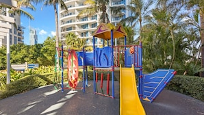 A playground with a yellow slide and climbing structures, surrounded by palm trees and modern buildings. - Chevron Renaissance Ocean View (Surfers Paradise)