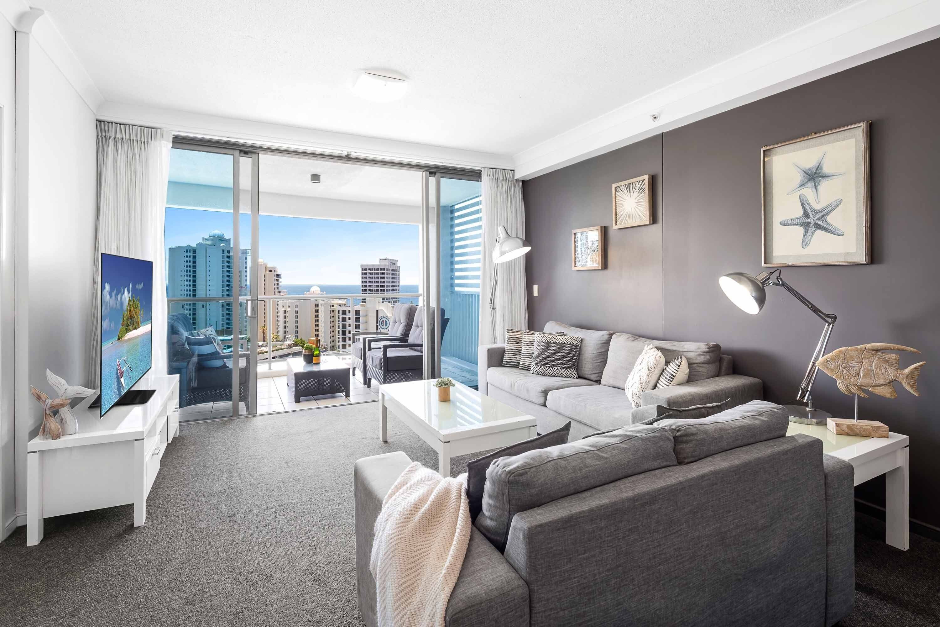 Una sala de estar moderna con un sofá gris, una mesa de centro blanca y un balcón con vistas al horizonte de la ciudad.