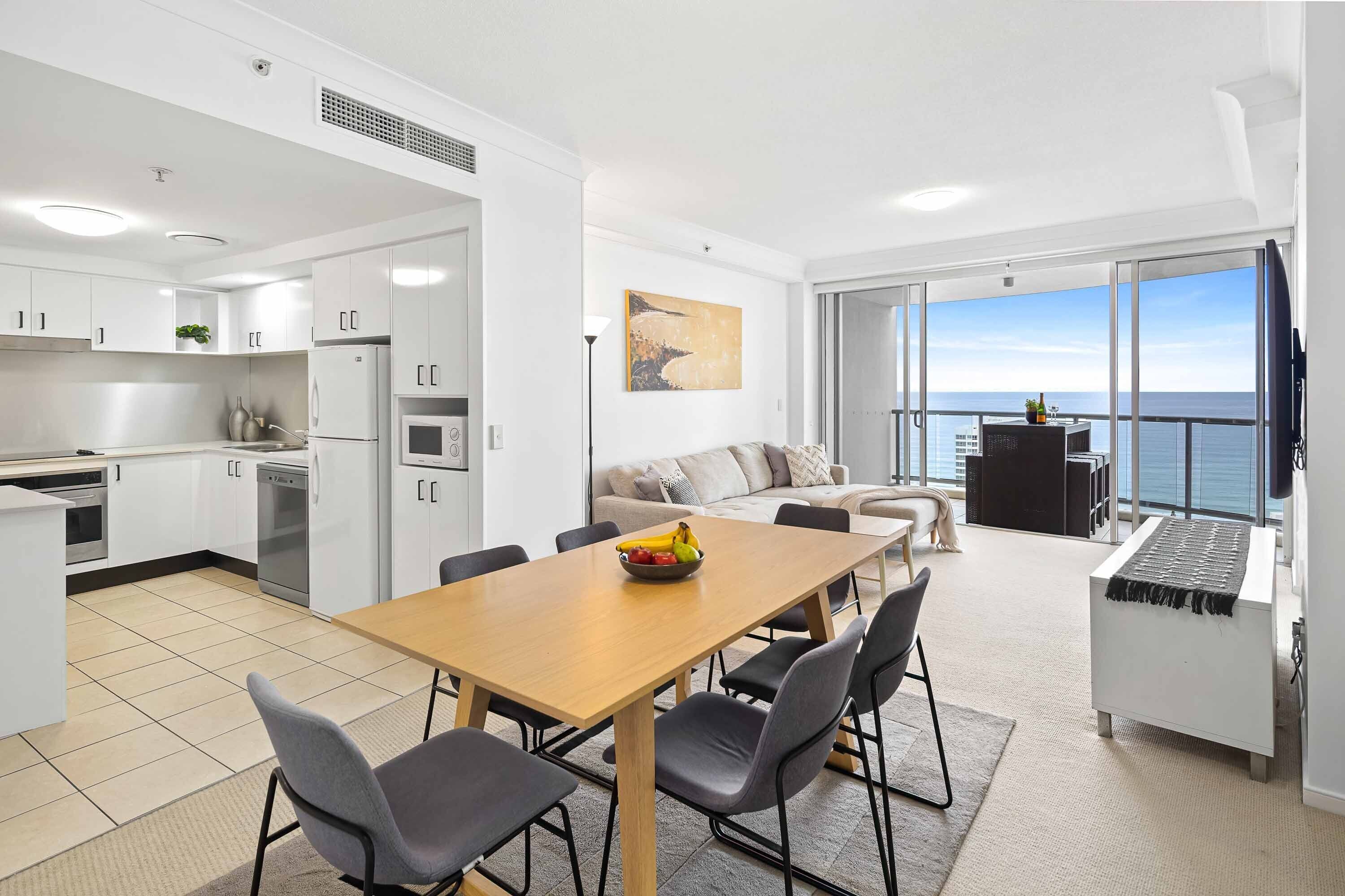 A modern kitchen with white cabinets, a dining area with a wooden table and chairs, a sofa, and a balcony with a view of the sea.