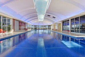 A large indoor swimming pool with clear glass walls, red chairs, and a ceiling with a blue light fixture.