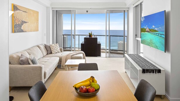 A modern living room with a balcony overlooking the sea, a dining table with fruit, and artwork on the walls. - Chevron Renaissance Ocean View (Surfers Paradise)