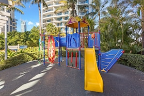 A playground with a yellow slide, blue and red play structures, and palm trees in the background.