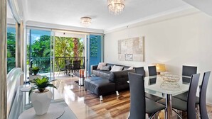A modern living room with a glass dining table, black leather sofa, and a balcony with a view of greenery. - Chevron Renaissance Garden View (Surfers Paradise)