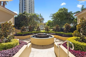 A landscaped courtyard with a central fountain, surrounded by well-maintained hedges and palm trees.
