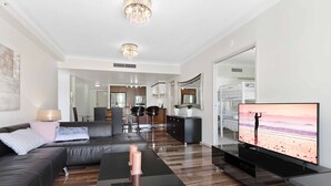 A modern living room with a leather sofa, a coffee table, and a flat-screen TV. There is a kitchen in the background with a dining area. - Chevron Renaissance Garden View (Surfers Paradise)
