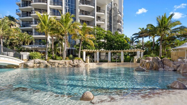 A clear pool with rocks, palm trees, and a building with balconies. - Chevron Renaissance Garden View (Surfers Paradise)