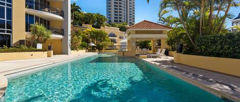 A swimming pool with a 1.2m depth warning sign, surrounded by a building with balconies and palm trees.