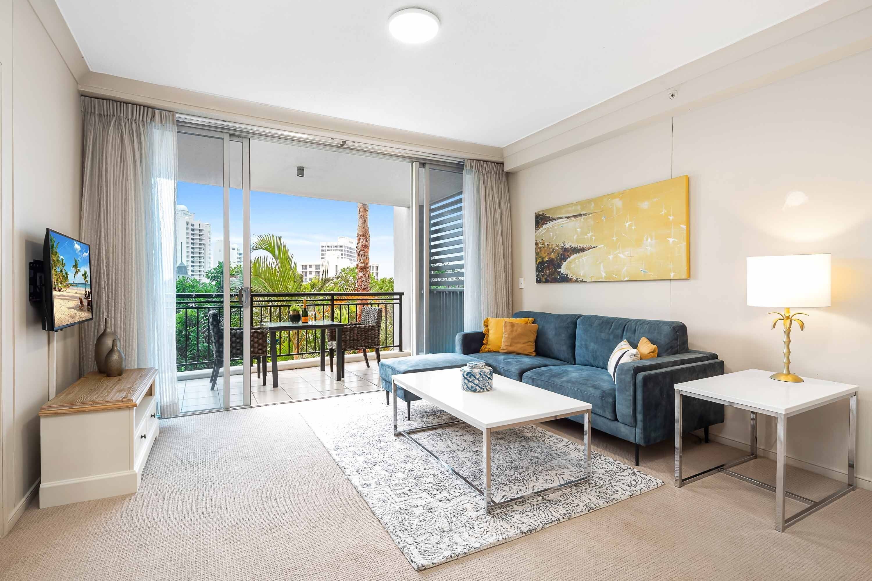 A modern living room with a sofa, coffee table, and TV, a balcony with a view of buildings and greenery.
