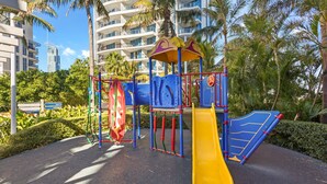 A playground with a yellow slide and climbing structures, surrounded by palm trees and modern buildings. - Chevron Renaissance Garden View (Surfers Paradise)