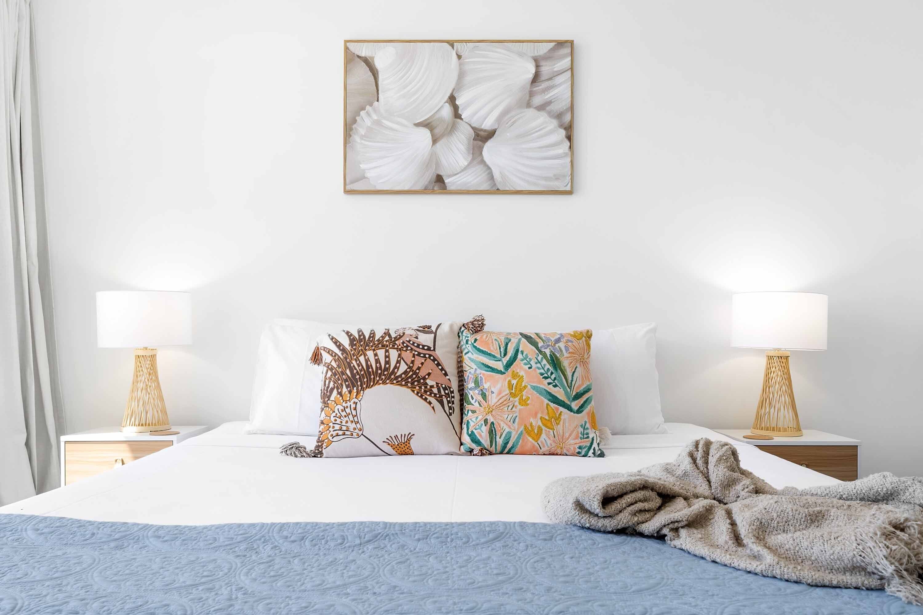 A neatly made bed with two pillows, a blue blanket, and a wall-mounted artwork of seashells.