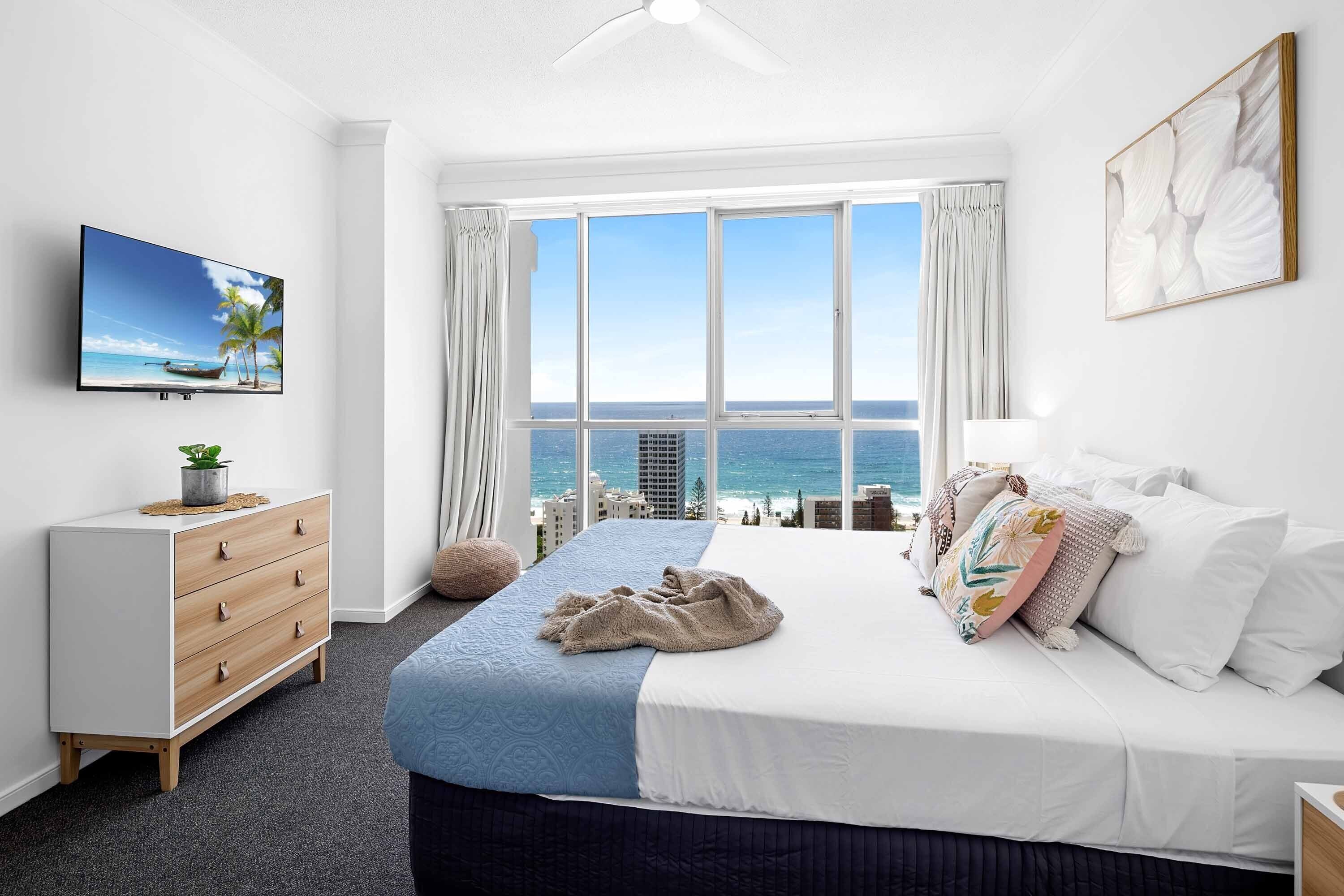 A bedroom with a bed, a dresser, a TV, and a large window with a view of the ocean.